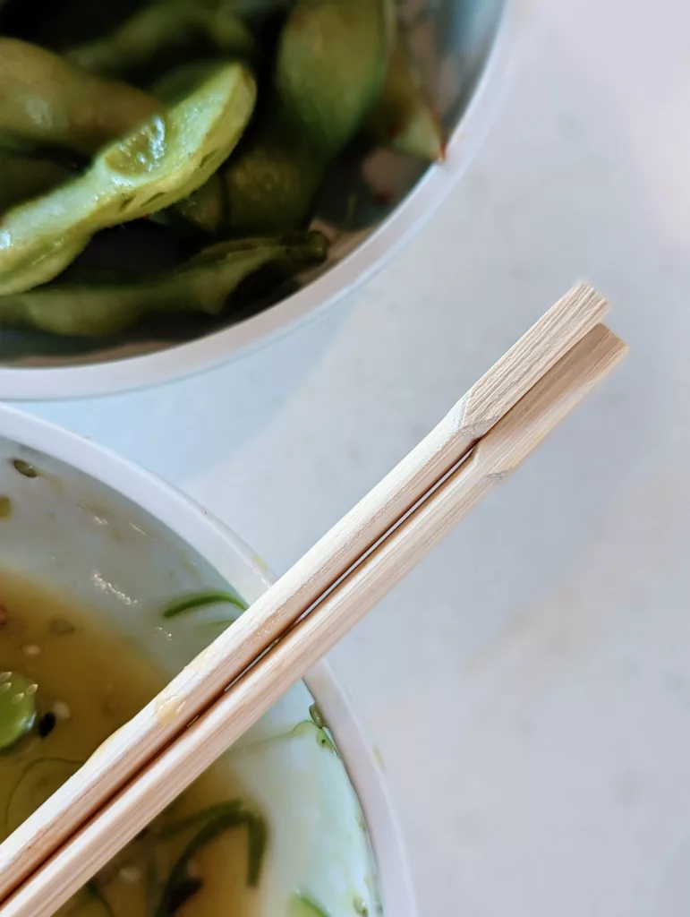 Wooden chopsticks resting over a bowl of clear broth beside a bowl of green edamame pods on a white table.